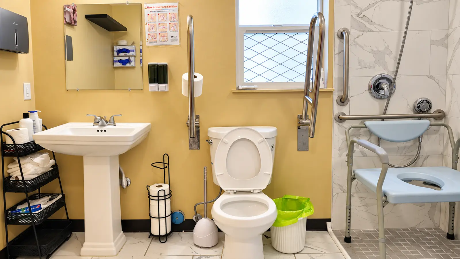 An accessible bathroom at Prime Adult Family Home with grab bars beside the toilet, a pedestal sink, and a roll-in shower with a shower chair