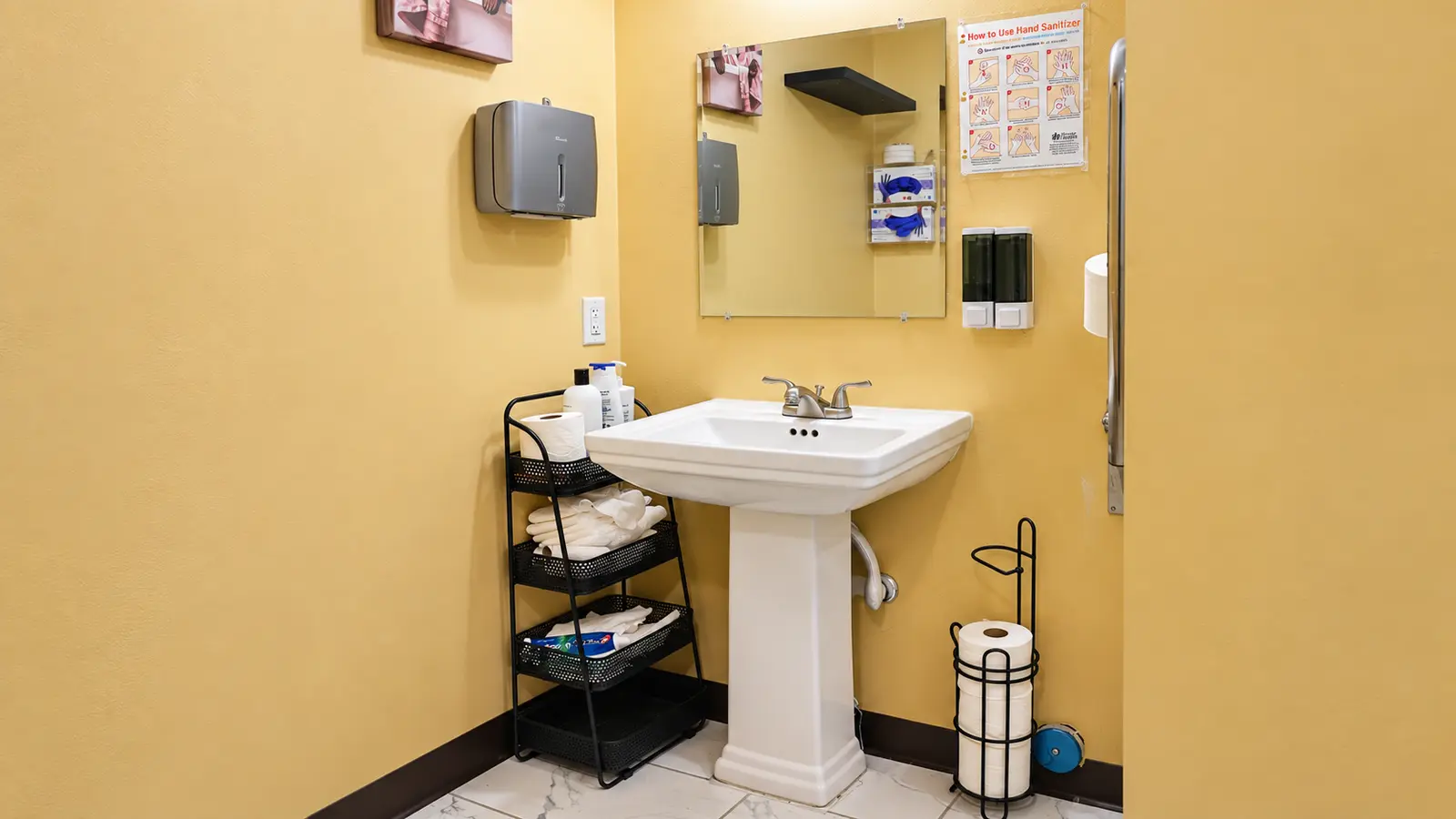 A pedestal sink and mirror at Prime Adult Family Home, with folded towels on an open shelf beside it